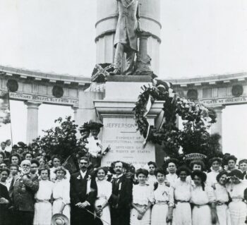 A large group of mostly women in white dresses and a few men in suits stand in front of the Jefferson Davis Monument on Monument Avenue in Richmond, Virginia. There is a large wreath hanging from the corner of the column the Davis sculpture stands upon.
