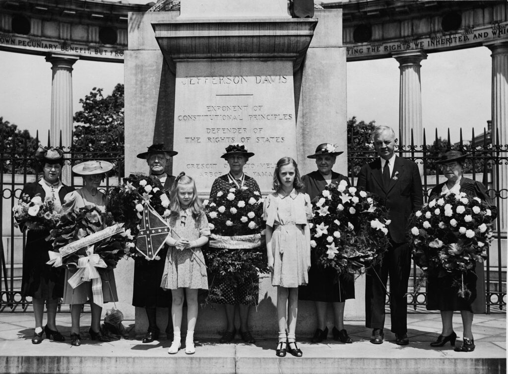 Six adult women wearing dresses and hats and one man in a suit with two young girls standing in front. The Jefferson Davis monument is behind them. The women are carrying large flower wreaths or a bouquet and one of the girls is carrying the Confederate flag.