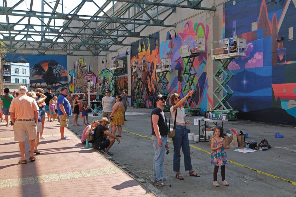 Shell of a building with two walls and only metal beams overhead. There are five artists painting murals and lots of folks watching.