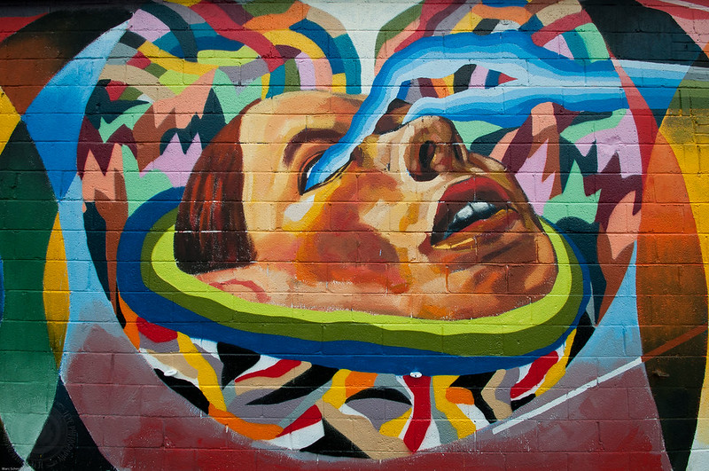 Mural of a face looking upward with blue streams of color coming out of the eyes on a colorful background.